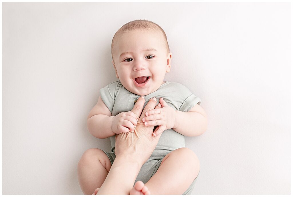 5 month old baby in sage green romper laying on back with a big grin on his face. Mom's hand is coming in from the bottom of the image and tickling his belly.