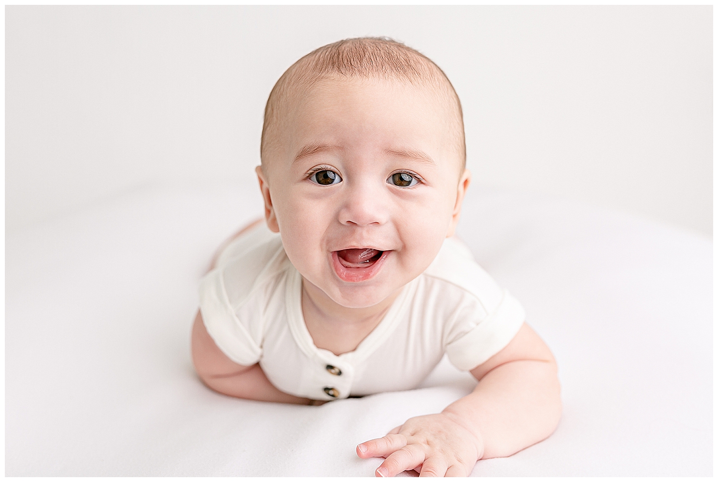 Baby with big brown eyes laying on belly holding up his head with a big gummy grin. Baby is about 6 months old and dressed in a white romper and laying on a white blanket.