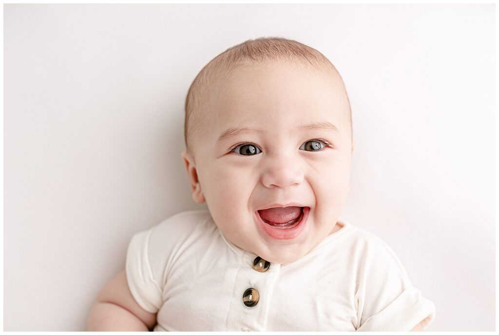 Five month old baby with brown eyes dressed in a white romper has a big smile on his face and is looking right at the camera.