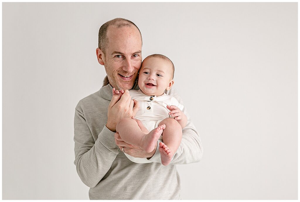 Dad dressed in light, neutral colors is holding 5 month old baby and their cheeks are touching. Baby is smiling off camera. Dad is smiling towards the camera.