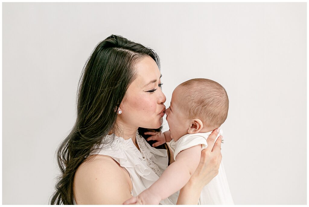 Mom is holding baby and kissing his nose. Mom has long dark hair and is dressed in white. Baby is also dressed in white.