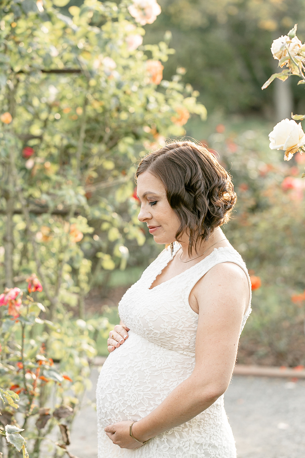 A pregnant woman in a white lace dress stands in a rose garden holding her bump