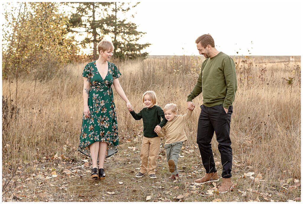 Family of 4 dressed in a green and beige color pallete walking outside in a field of golden grass. They are holding hands and laughing with each other at their fall family portraits.