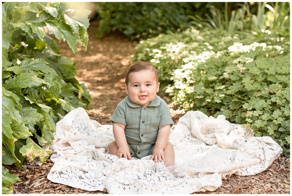 6 month old baby boy dressed in sage green romper with buttons down the front and a little collar. He is sitting on a vintage crochet blanket surrounded by greenery and smiling at the family photographer.