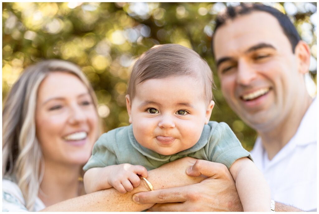 Mom and dad are holding baby boy having him lying on his belly. Baby is in focus and the parents are a bit blurred behind him and they are smiling looking at the baby. Baby has his tongue sticking out and is looking at the family photographer.