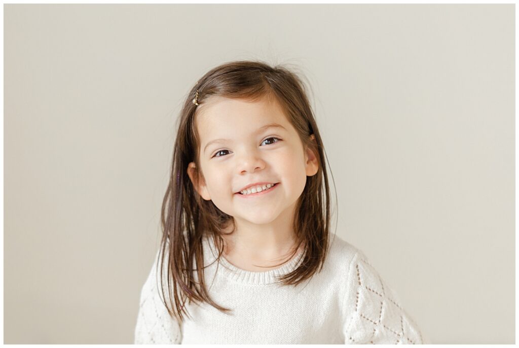 Toddler girl in white dress and dark brown hair smiling at the camera.