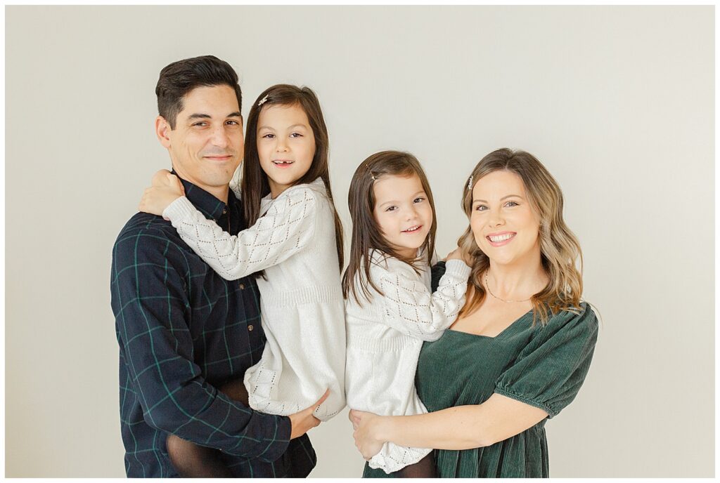 Family of four Dad, two daughters and Mom all being snuggly and looking at the camera and smiling. They are dressed in whites, blues, and greens. Family Studio Mini Photo Session.