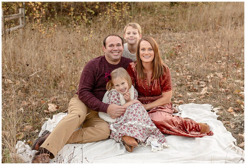 family of four sitting down on a blanket among the fall leaves at their fall family photography session.