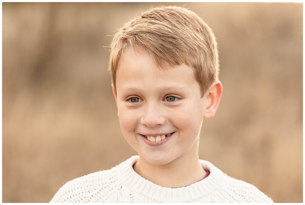 Tight shot of a young boy with blonde hair wearing a cream sweater and smiling slightly off camera. 