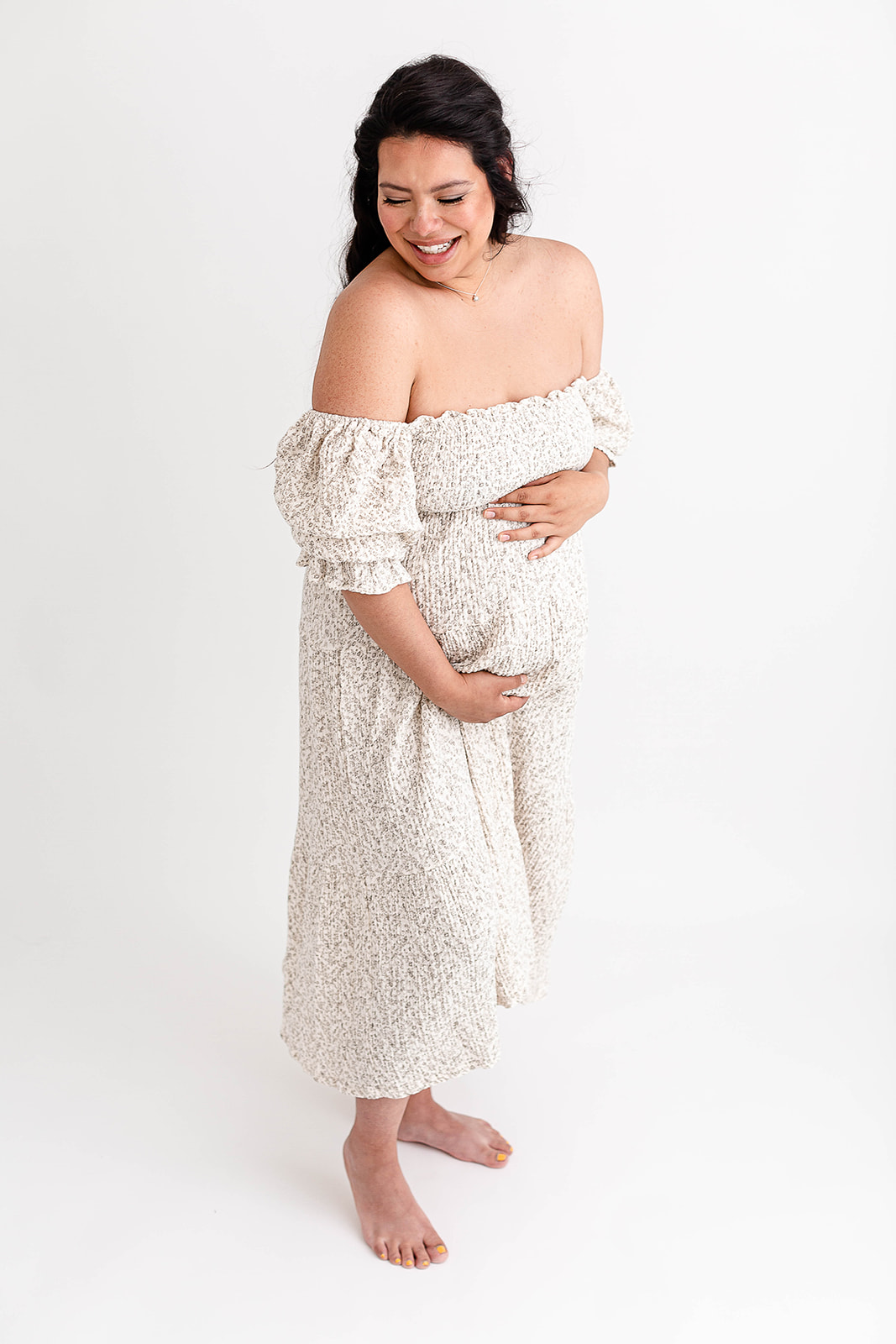 A mother to be laughs down her shoulder while standing in a studio holding her bump