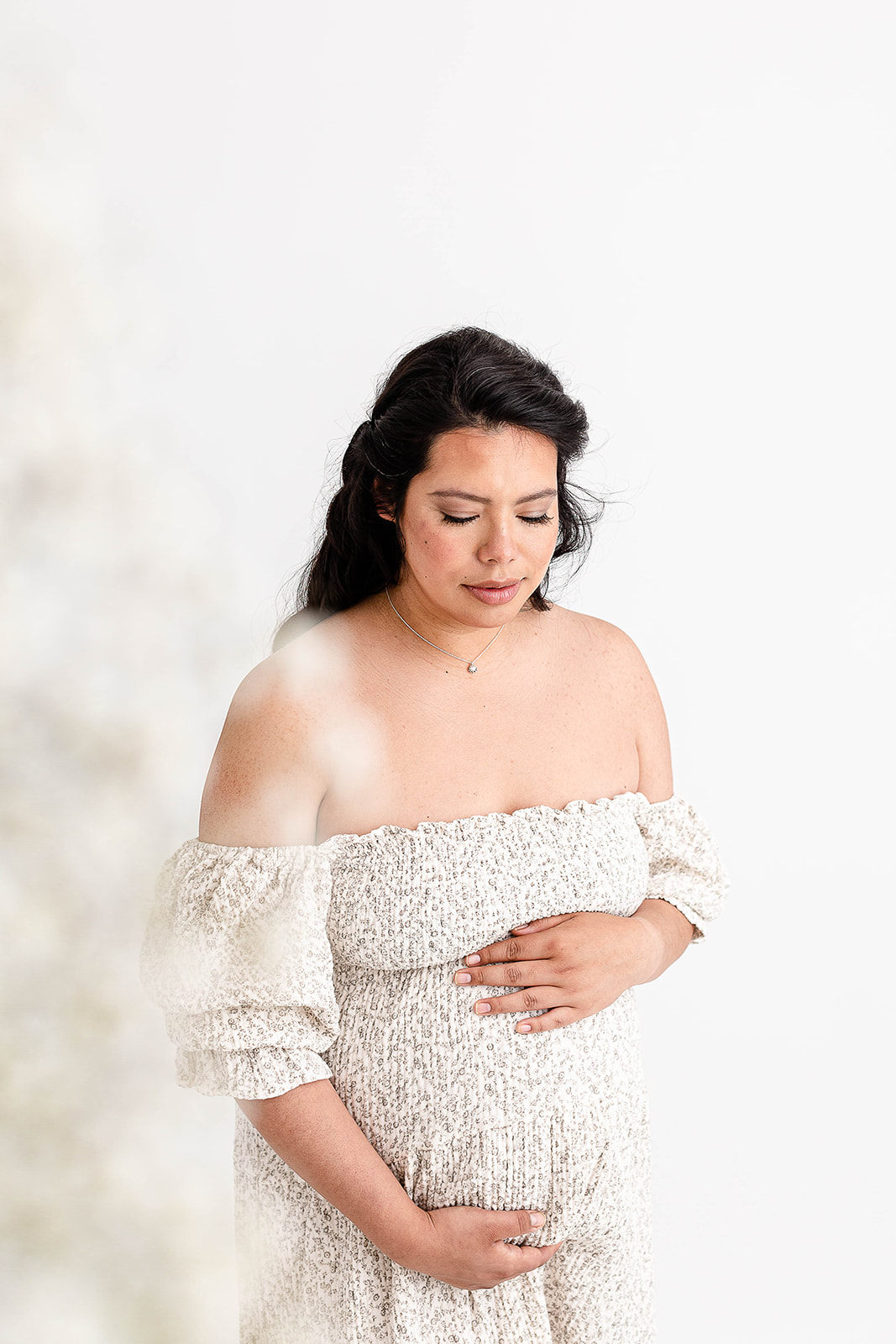 A mom to be stands in a studio smiling down to her bump in a white patterned dress before calling a Portland Lactation Consultant