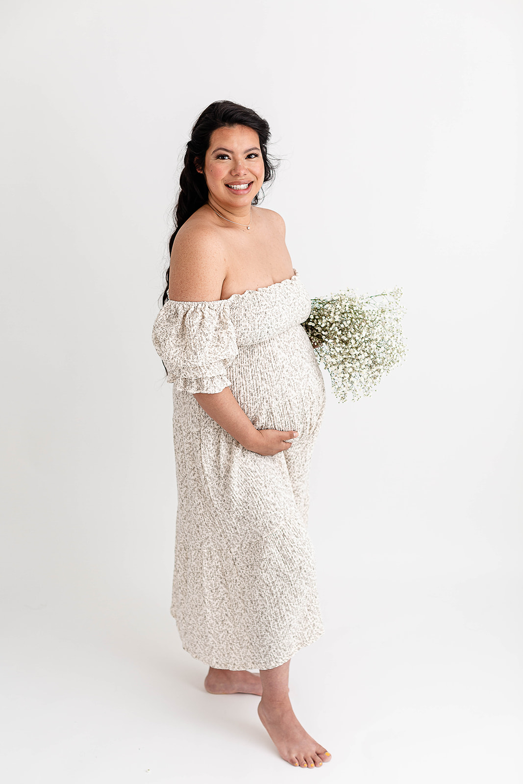 A mom to be in a white maternity dress stands in a studio holding a bouquet of white flowers and her bump before calling a Portland Lactation Consultant
