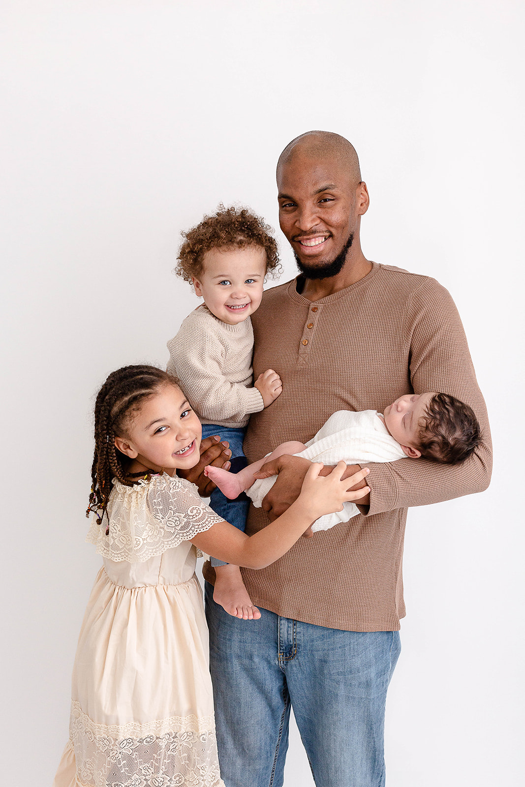 A father in a brown shirt stands in a studio holding his newborn baby and young toddler in each arm while his older daughter hugs them all because they use Portland Nannies