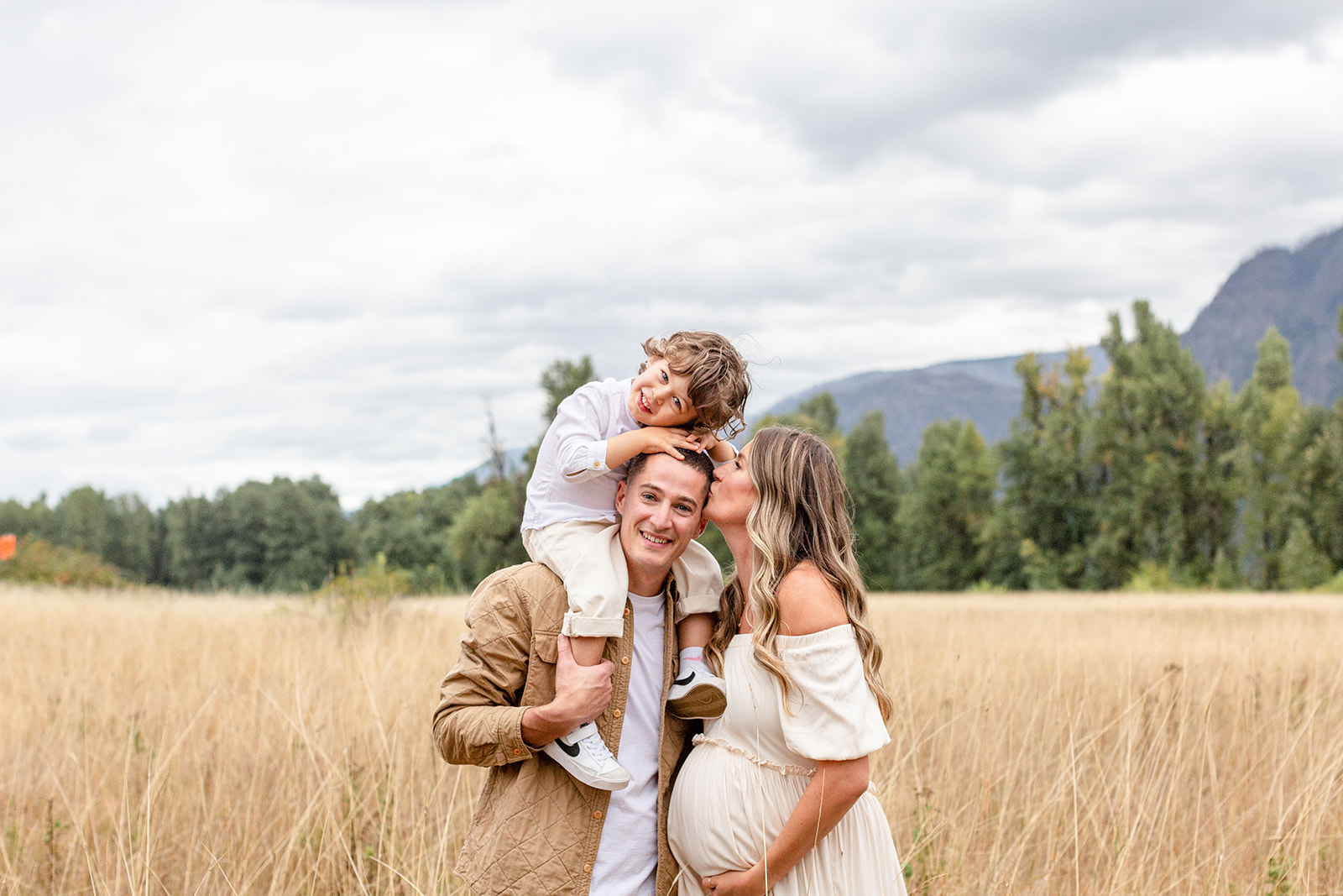A pregnant mom kisses dad's head while their young son sits on his shoulders in a field of tall grass with help from prenatal chiropractor portland