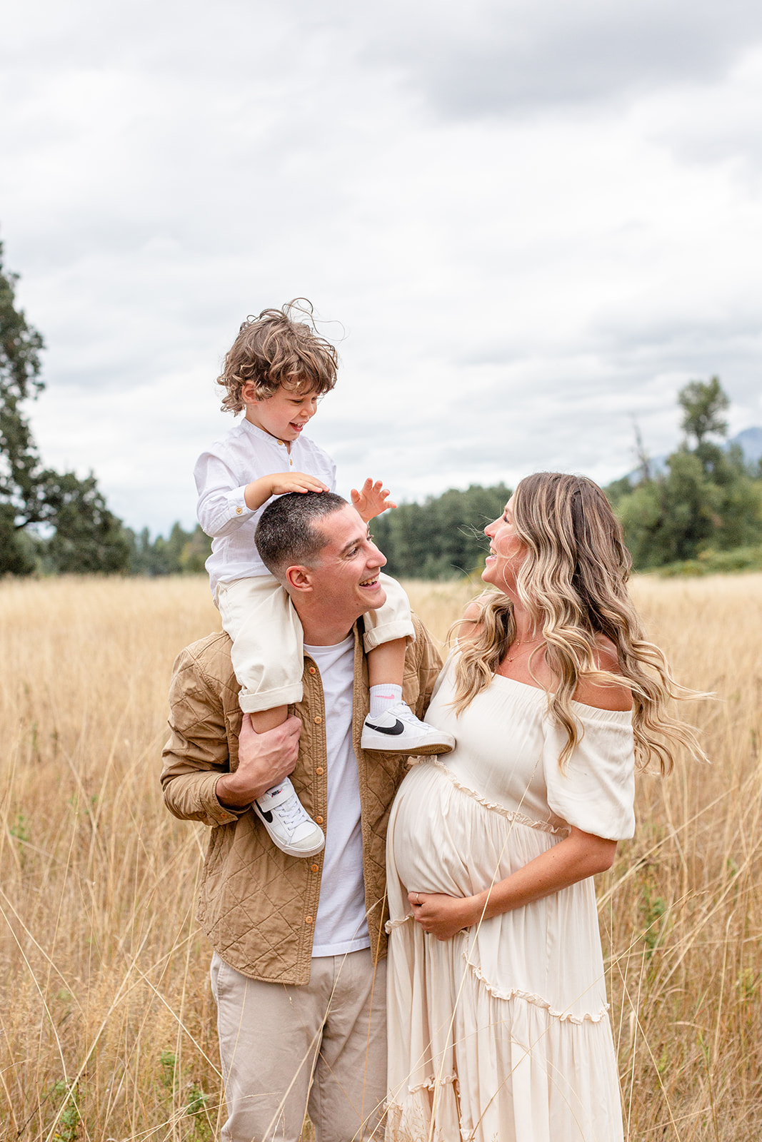 A young boy sits on dads shoulders while mom laughs with them and holds her pregnant bump in a field of tall golden grass thanks to a prenatal chiropractor portland