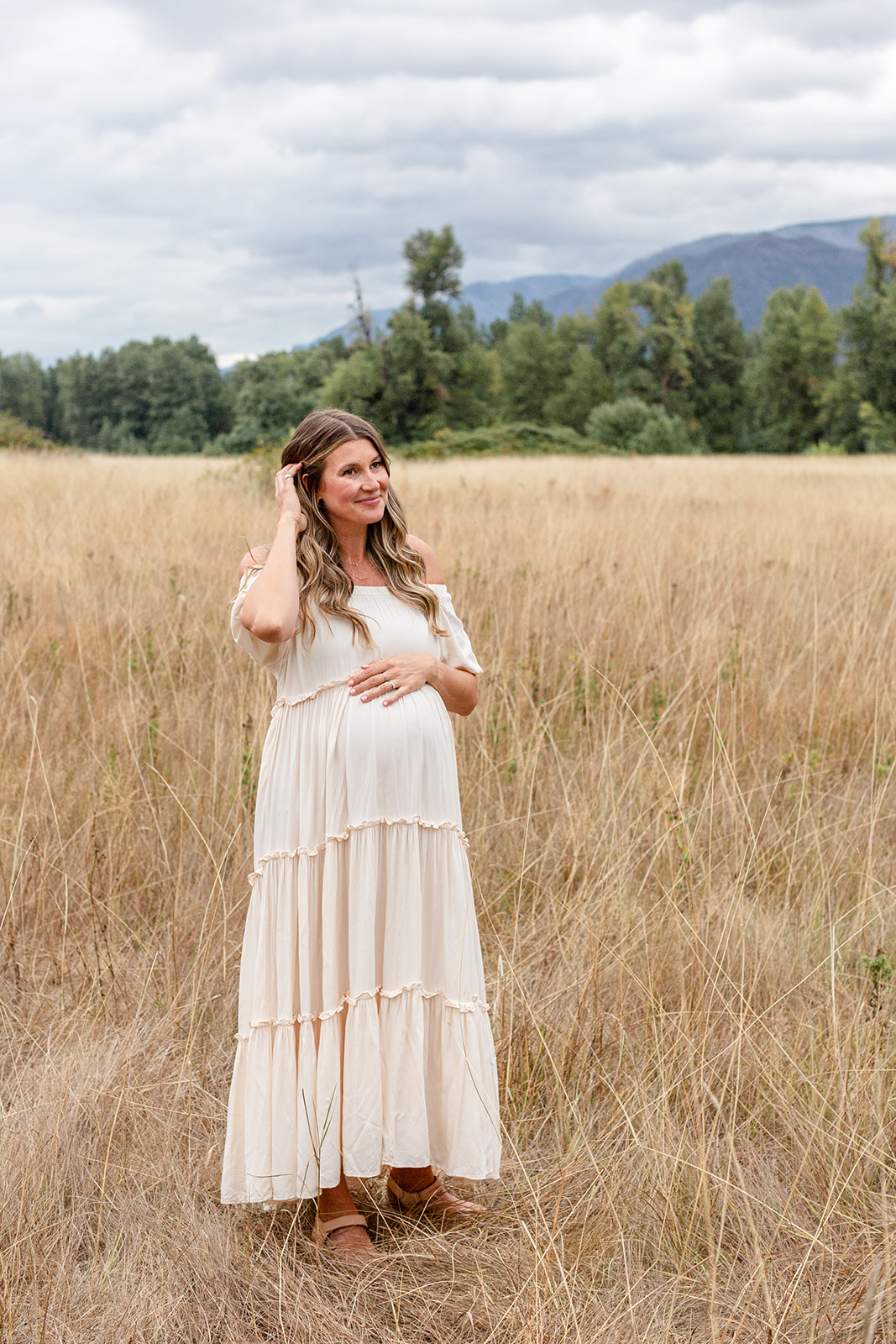 A mother to be in a white maternity gown stands in a field of tall golden grass pulling her hair back