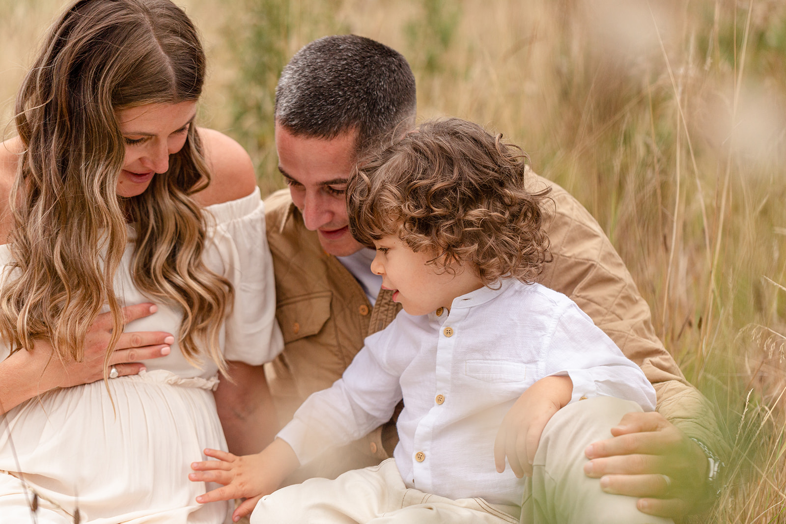 A pregnant mom sits in some tall grass with her husband and toddler son touching the bump