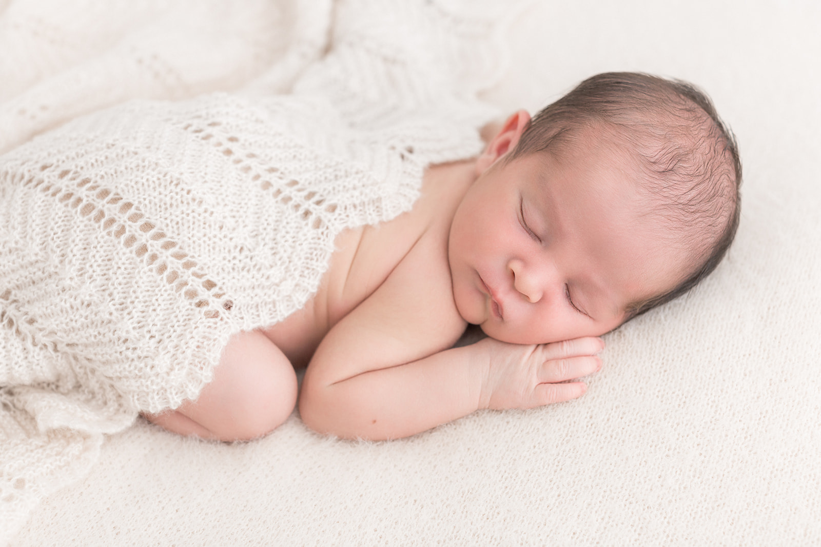 A newborn baby sleeps under a knit blanket on a white bed thanks to Portland Fertility Clinics