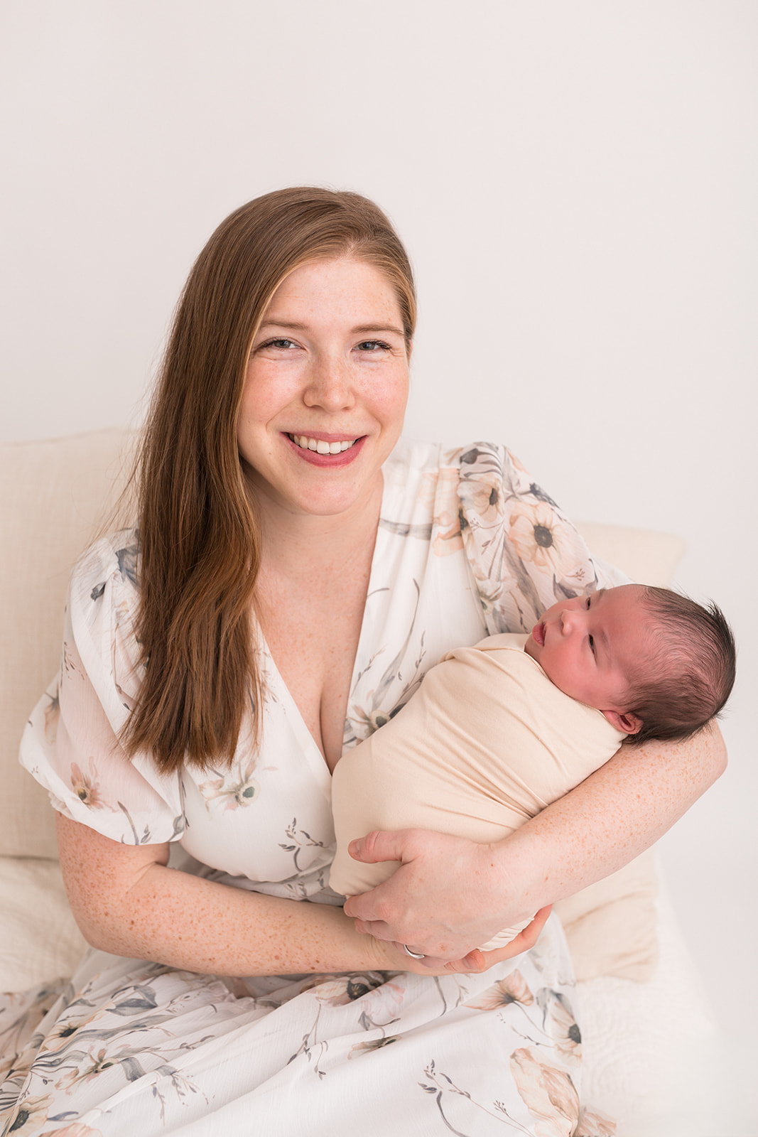 A mother in a floral dress sits on a bed smiling with her newborn baby in her arms