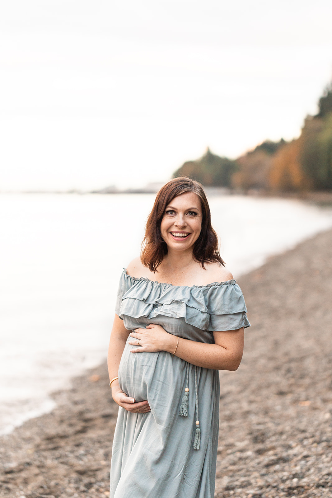 A mother to be in a blue maternity dress stands on a beach holding her bump