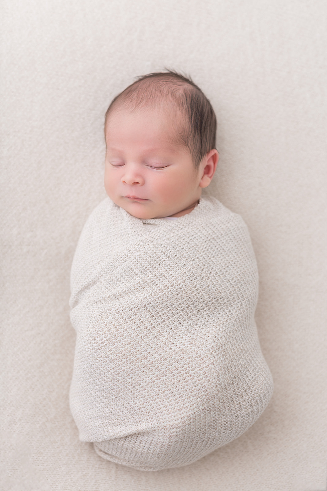 A newborn baby in a white knit swaddle sleeps on a white bed in a studio