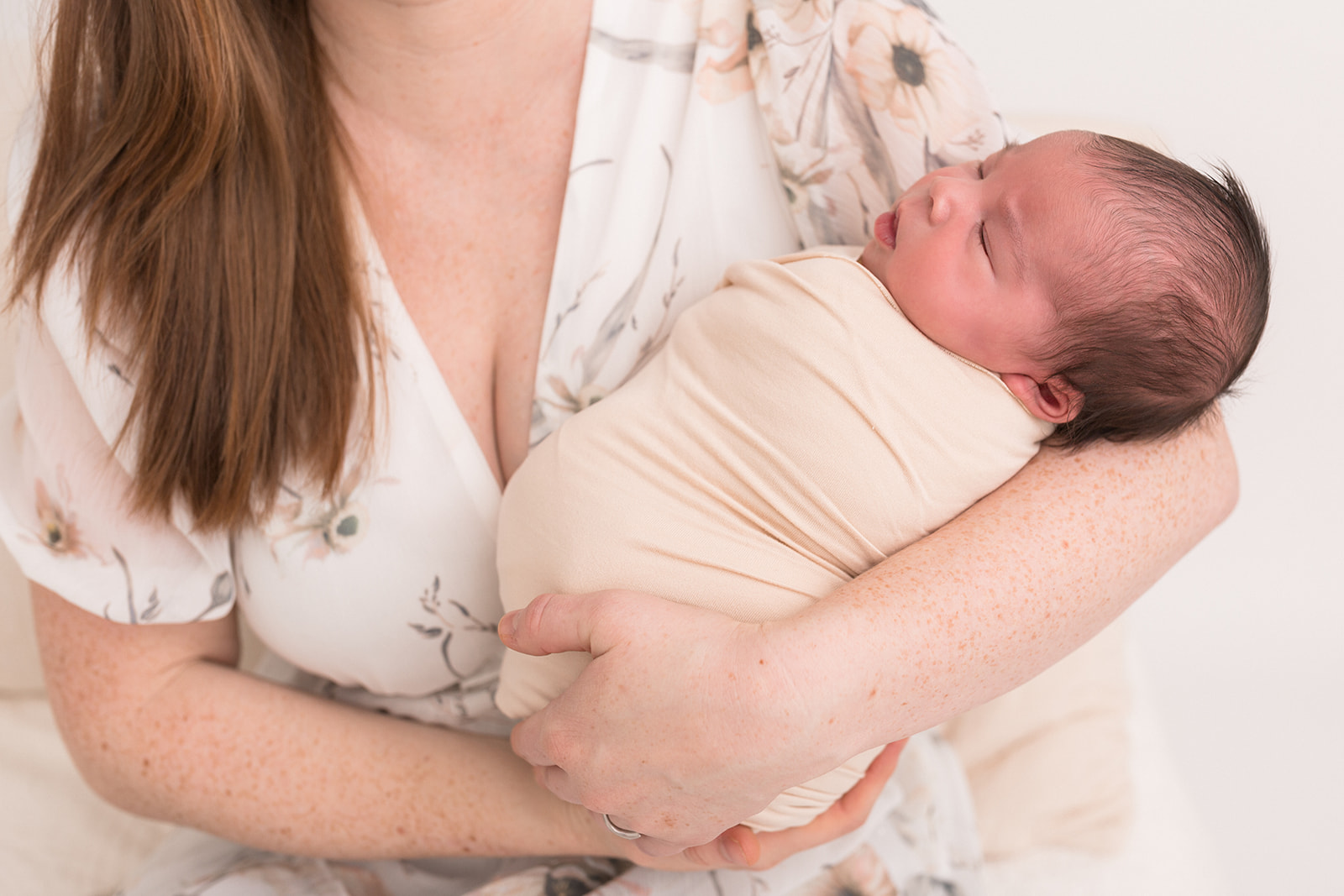 A newborn baby sleeps in mom's arms while she sits on a chair in a studio