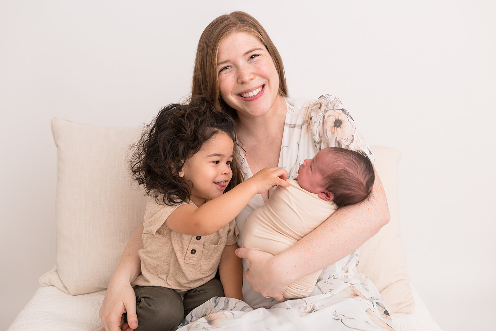 A happy mother sits on a chair in a studio playing with her toddler son and newborn baby daughter in her lap