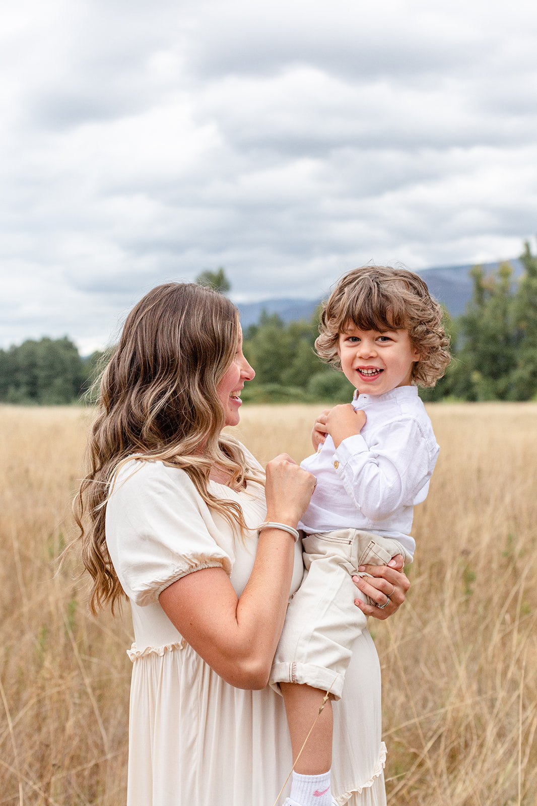 A toddler boy in khakis and a white button down sits on mom's pregnant bump while standing in a field of tall golden grass