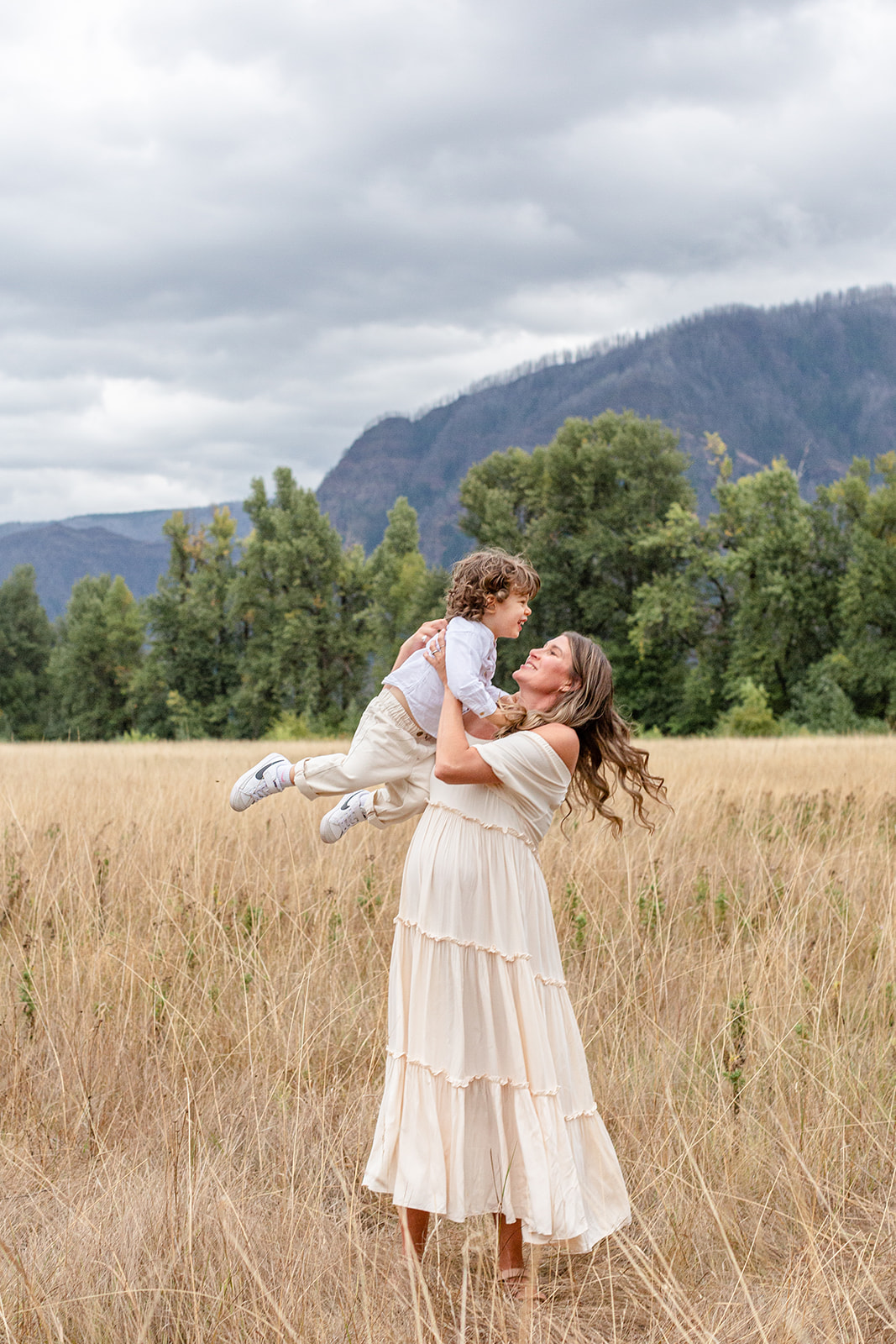 A mother in a white dress spins and dances with her toddler son in a field of tall golden grass after visiting a Portland Pediatric Dentistry