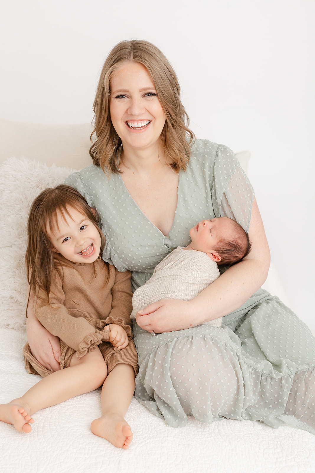A happy mother laughs with her toddler daughter while sitting on a bed in a studio as her newborn baby sleeps in her arms