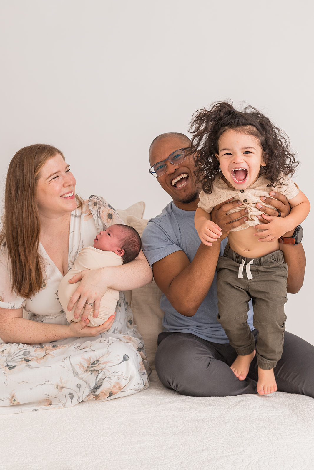 Happy parents play with their toddler and newborn baby while sitting on a bed in a studio