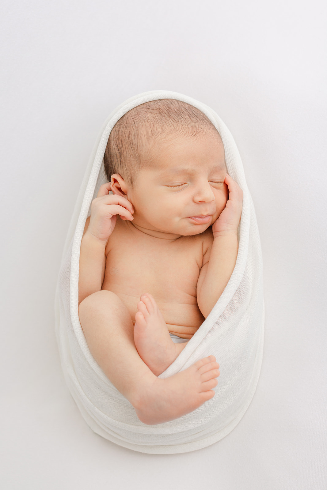 A newborn baby sleeps in a white open swaddle in a studio with hands on its head before some baby classes in Portland