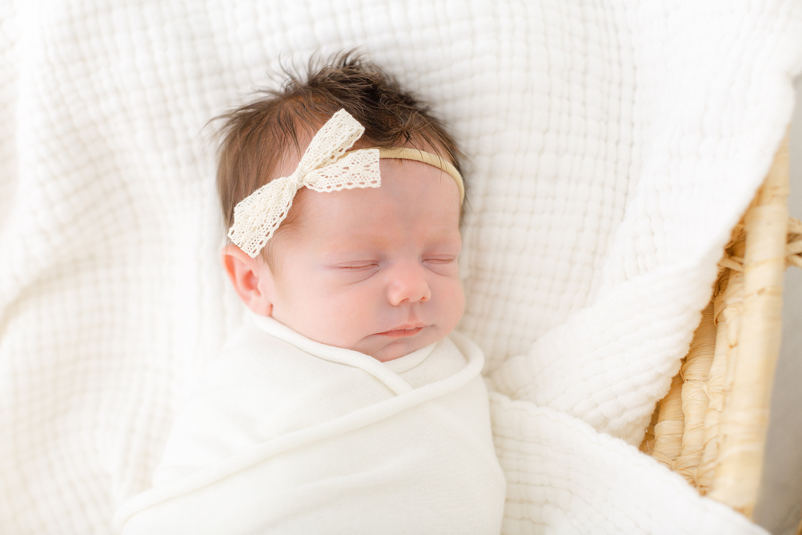 A newborn baby girl sleeps in a woven basket in a white swaddle