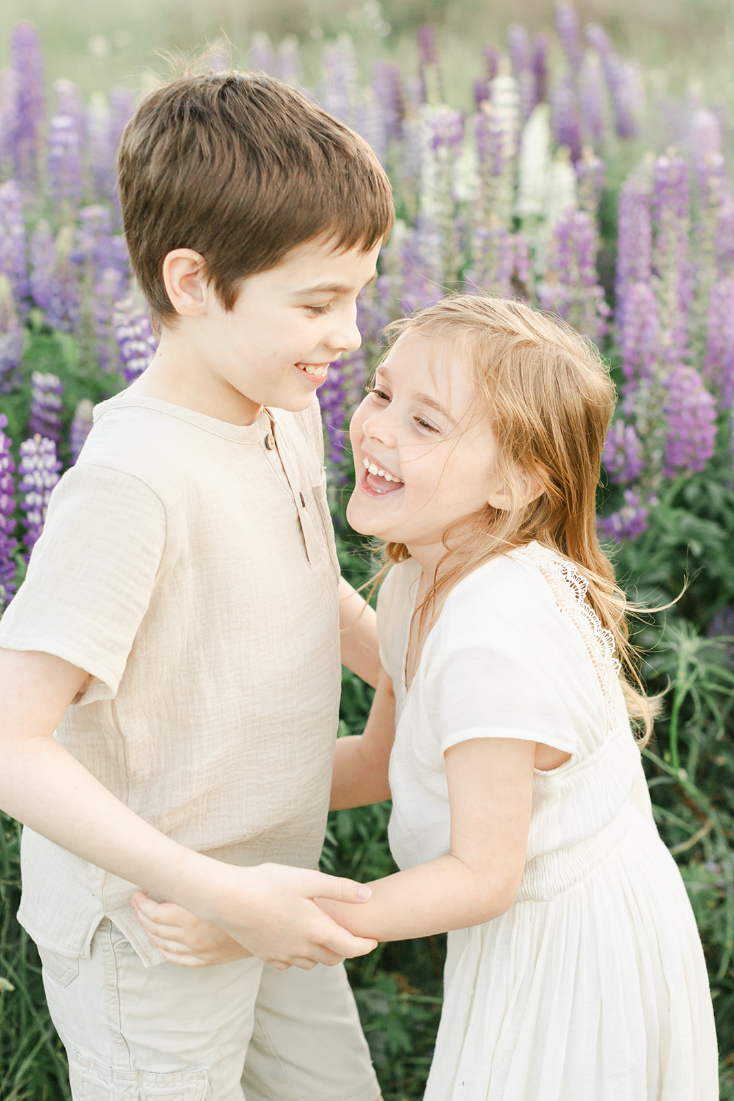 Young brother and sister hug and laugh while playing in a field of purple wildflowers