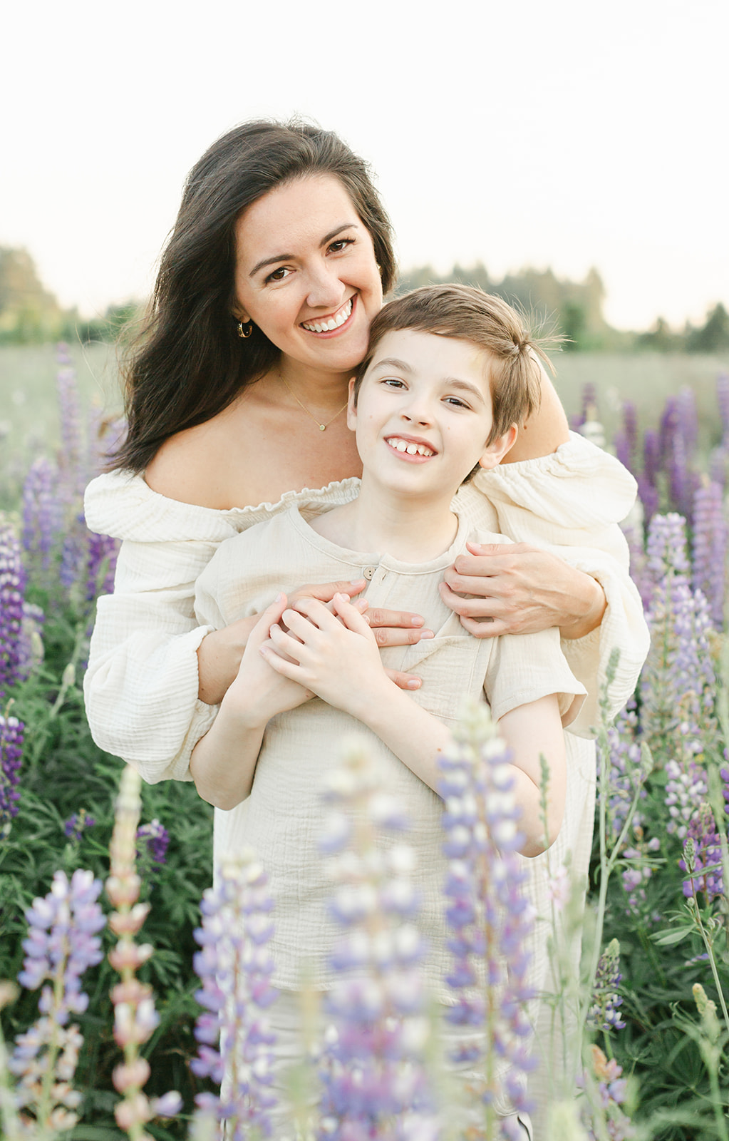 A mom in a white dress hugs on her smiling young son while standing in a field of purple wildflowers after visiting Portland Toy Stores