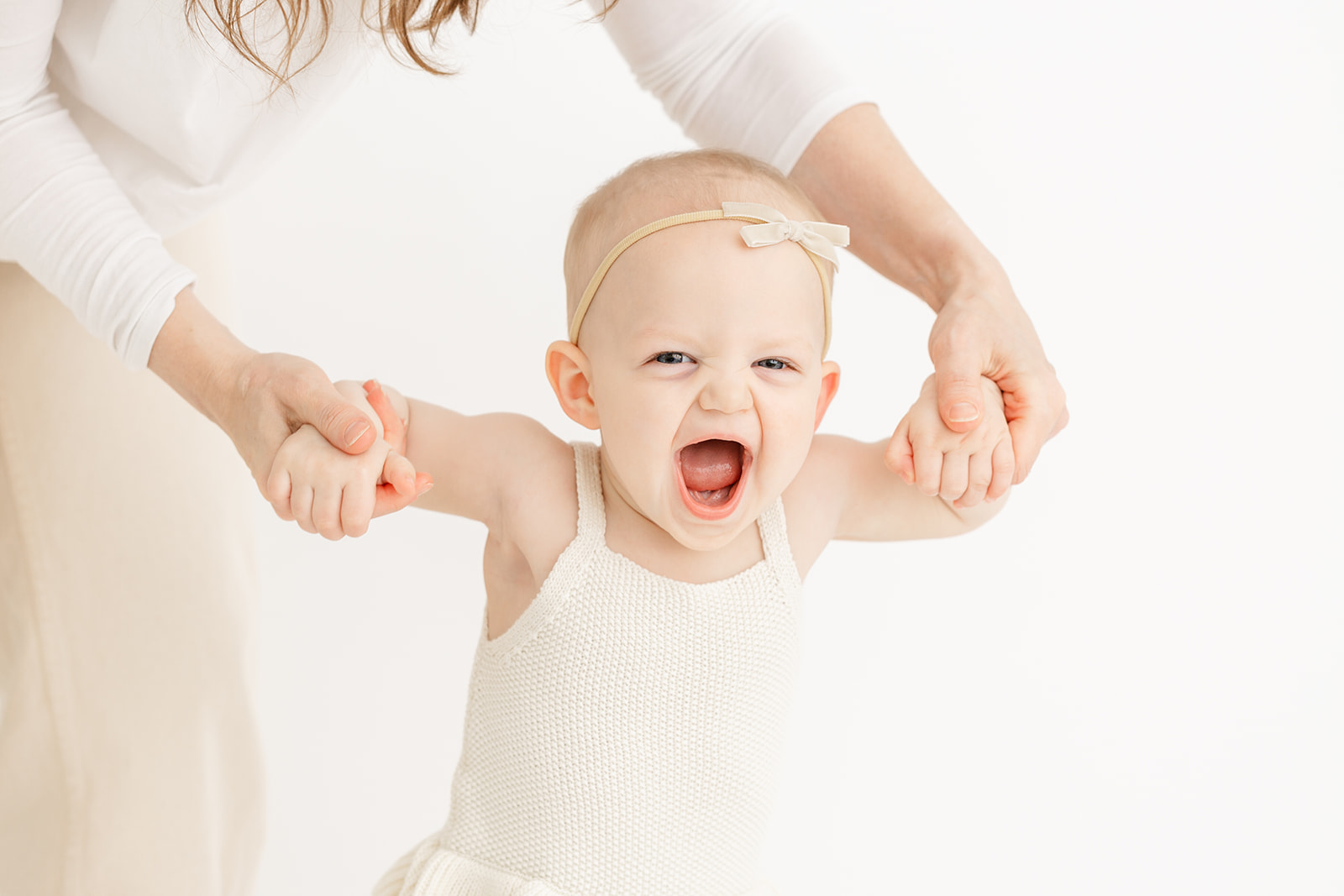 A mother helps her laughing infant daughter walk through a studio in a white dress
