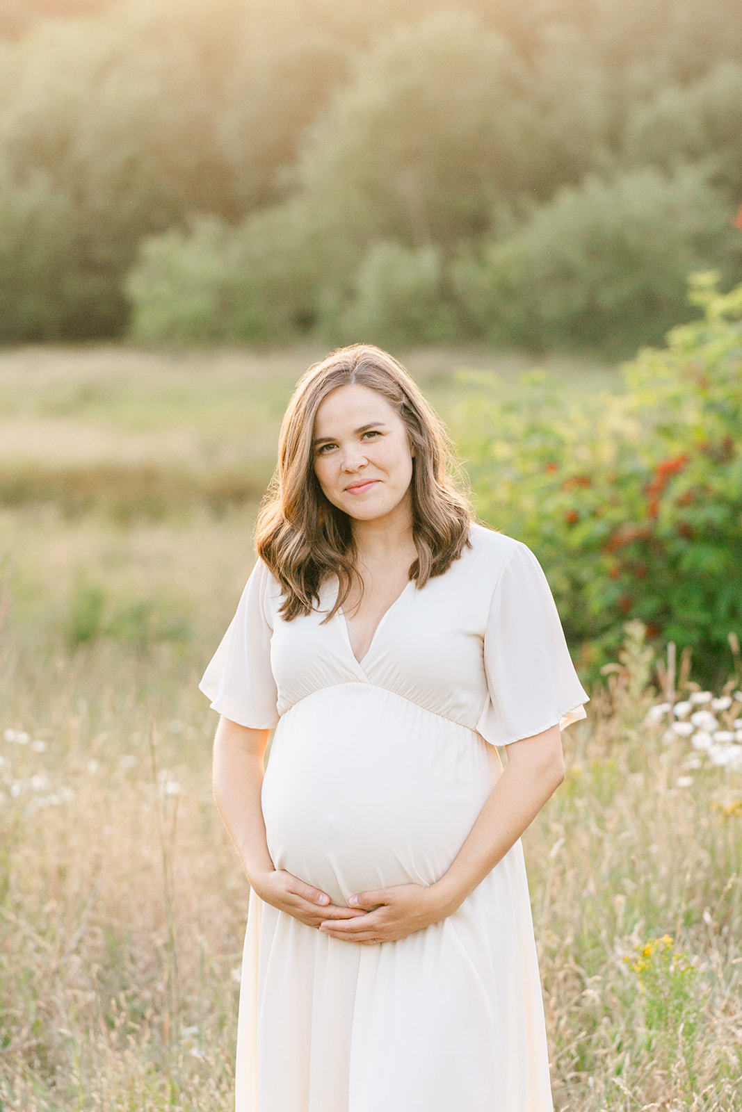 A pregnant mother to be smiles while holding her bump in a white maternity gown in a field of wildflowers at susnet after visiting Birth First Doulas