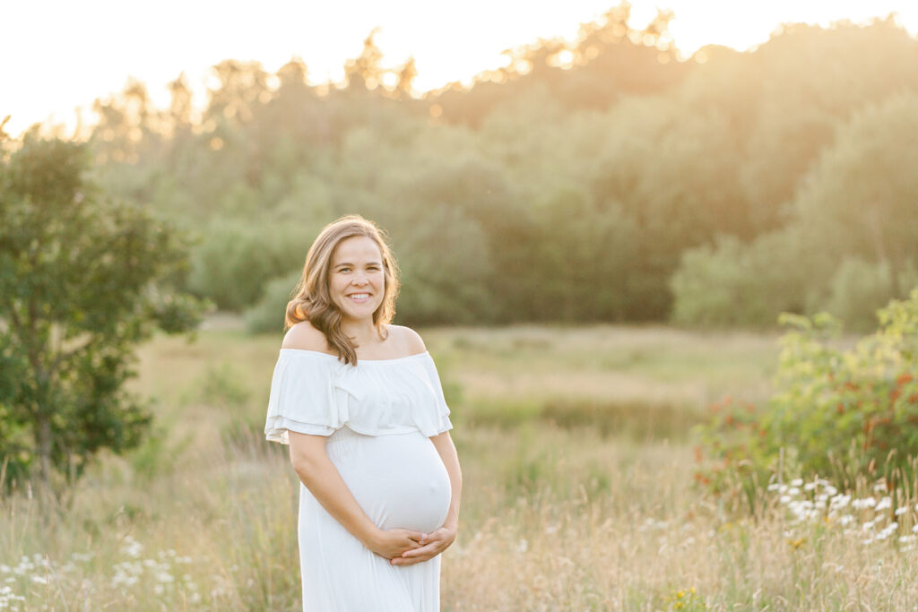 A mother to be stands in a field of wildflowers at sunset in a white maternity gown