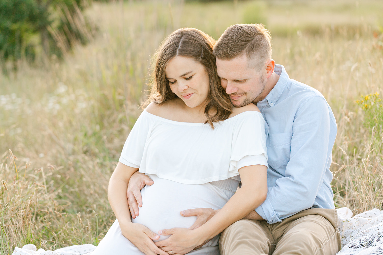 A pregnant couple sits in a field of tall glass snuggling while holding the bump on a picnic blanket at sunset after visiting Brave Birth
