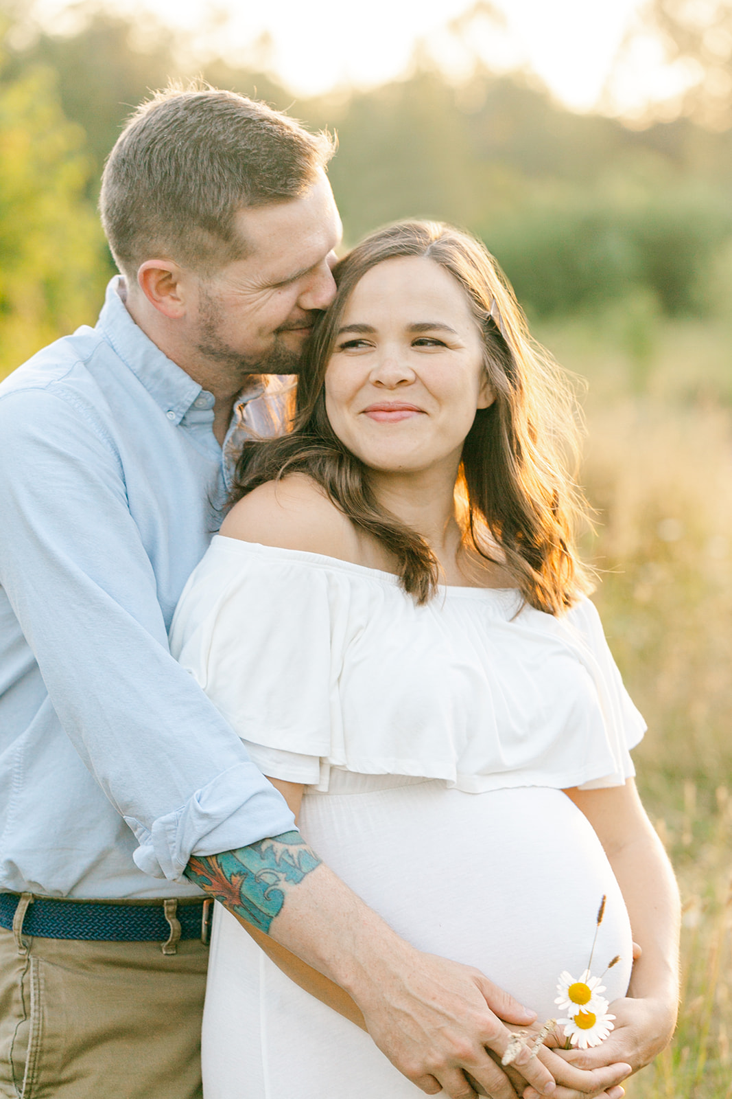 Happy expecting parents snuggle while exploring wildflowers in a park at sunset after meeting Brave Birth