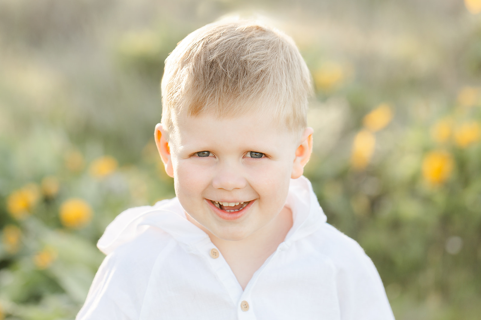 A toddler boy in a white shirt smiles big in a park with wildflowers at sunset