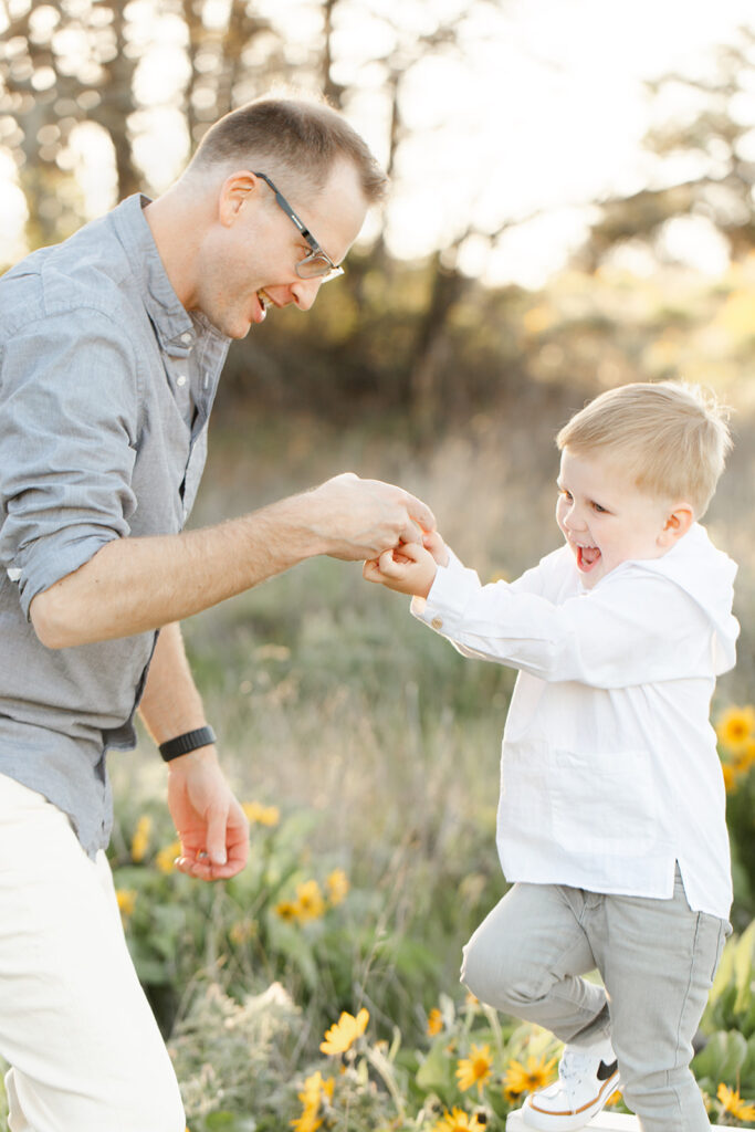 A happy toddler boy in a white shirt plays with dad while exploring a park at sunset after visiting Portland Preschools