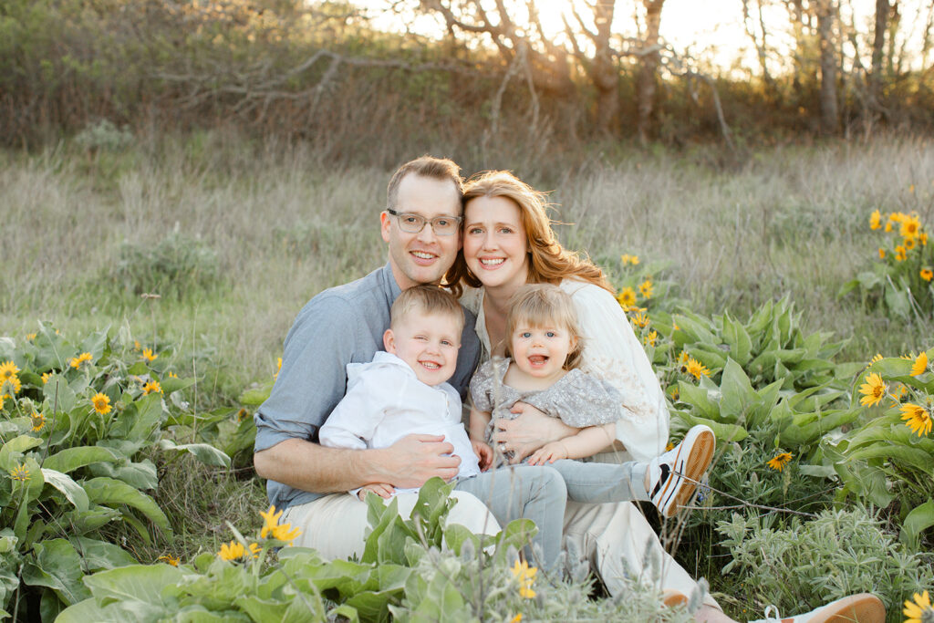 Happy parents sit among wildflowers on a windy day with their laughing toddler son and daughter in their laps after visiting Portland Preschools