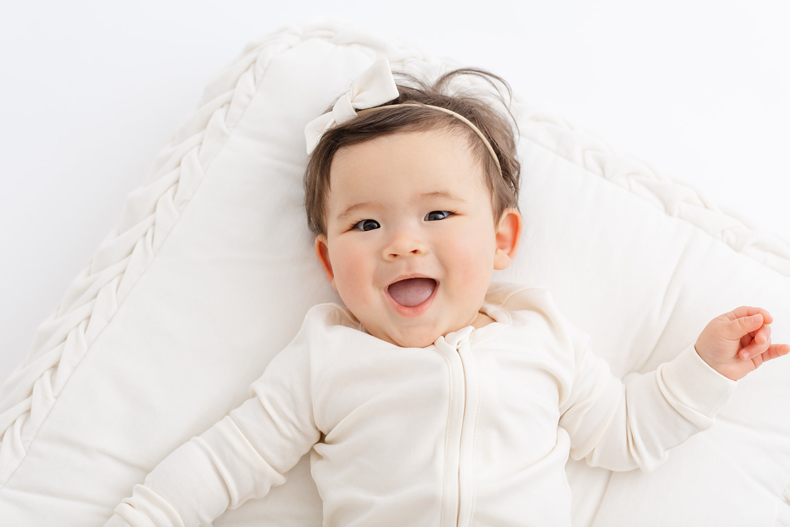 A newborn baby laughs while laying on a white pillow in a white onesie and bow thanks to Alma Midwifery