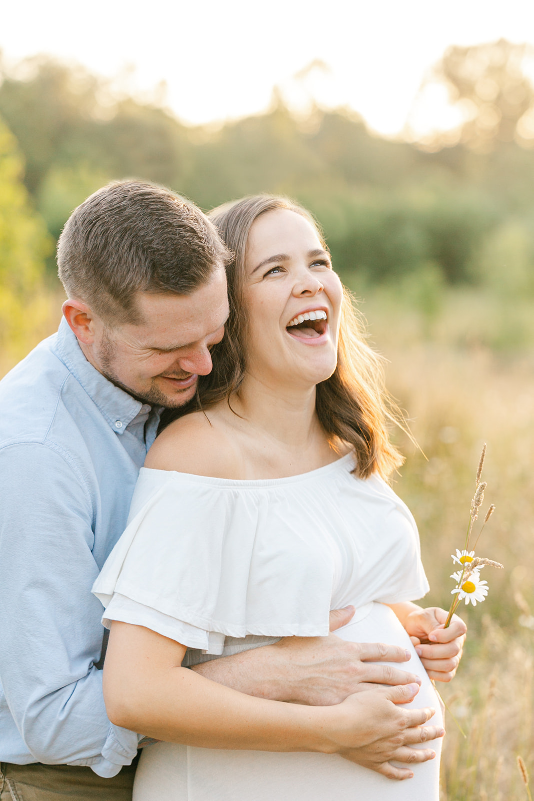 A mom to be laughs while being hugged around the bump by her husband