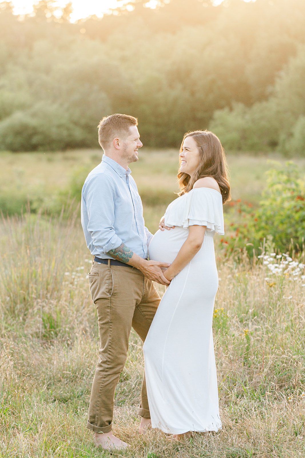 Happy expecting parents hold the bump while standing in a field of wildflowers at sunset after visiting Hearth and Home Midwifery