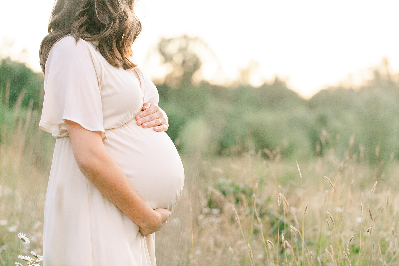 Details of a mom to be holding her bump in a white maternity gown in a field of tall grass after meeting Hearth and Home Midwifery