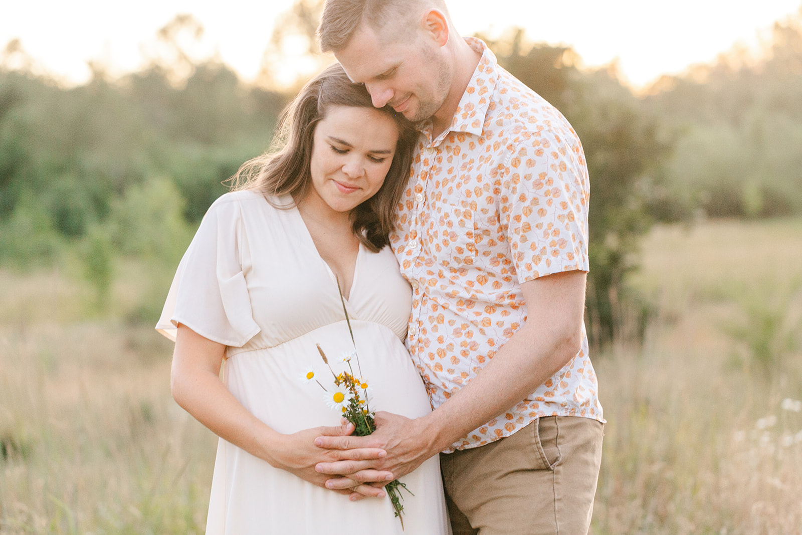 Expecting parents smile while holding the bump in a field of tall grass at sunset