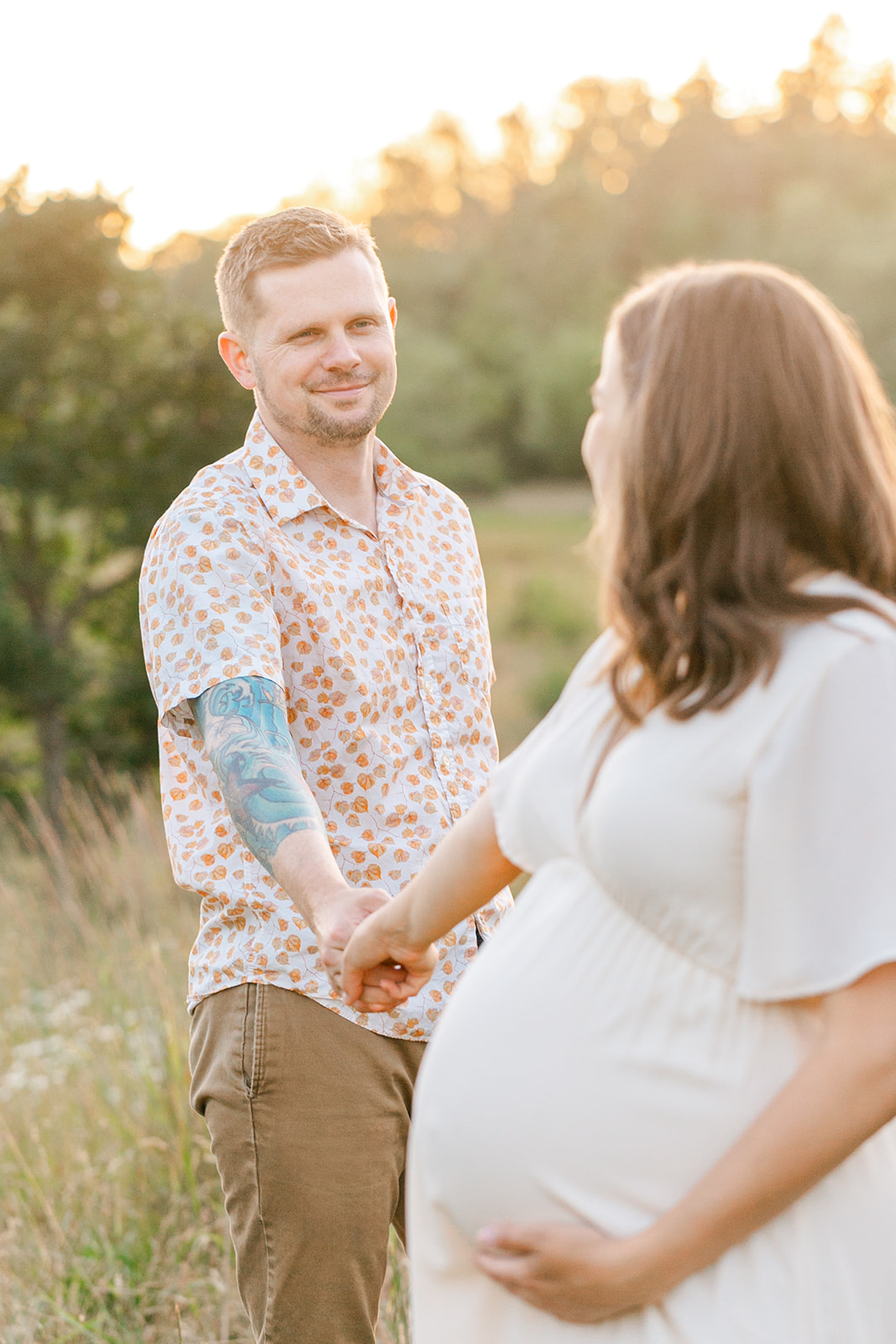 A mom to be in a white maternity dress leads her husband through a park trail at sunset by the hand after meeting Rosehip Midwifery