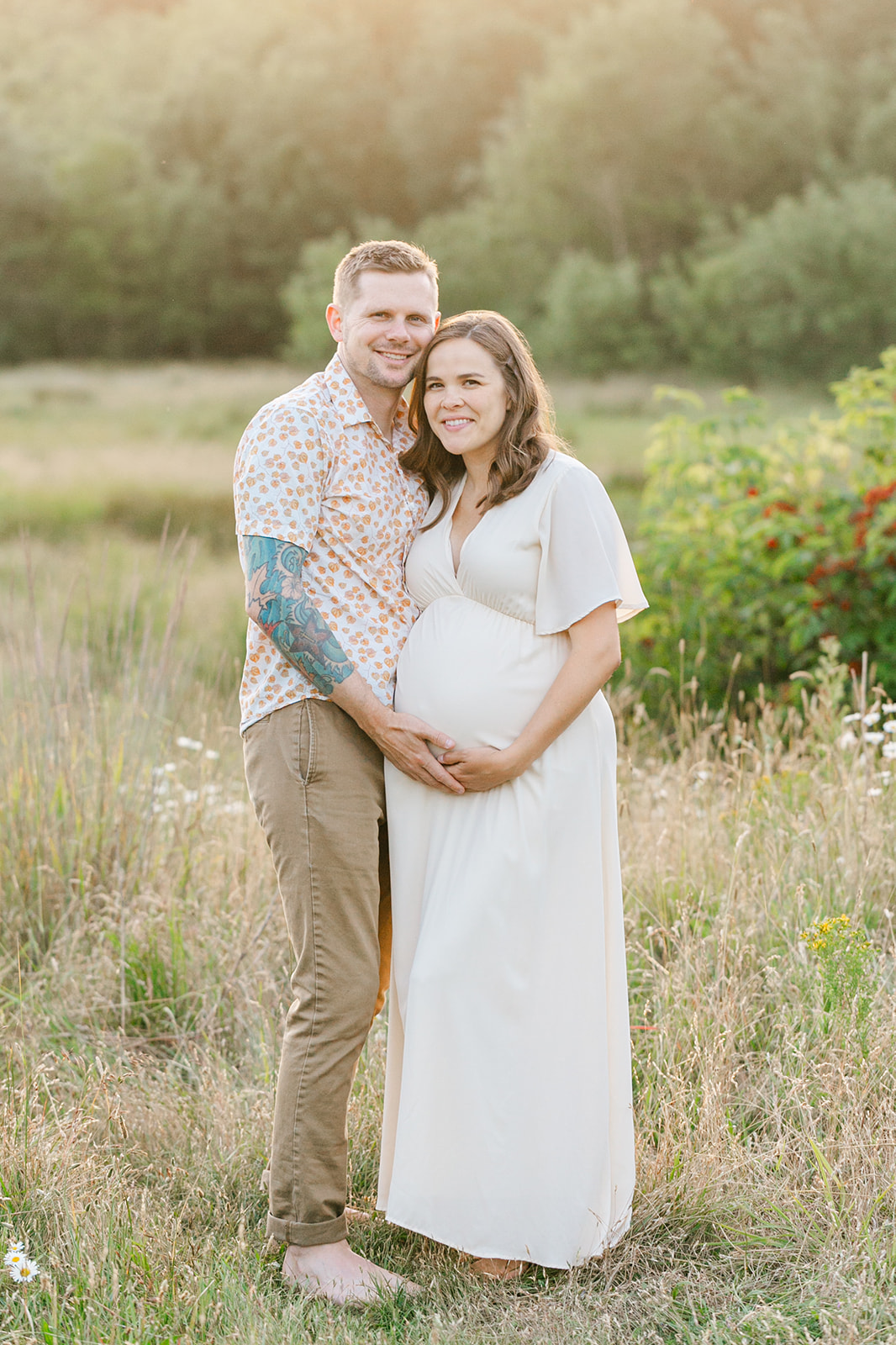 Happy expecting parents stand in a field of tall grasses and wildflowers holding the bump and touching cheeks after visiting Rosehip Midwifery